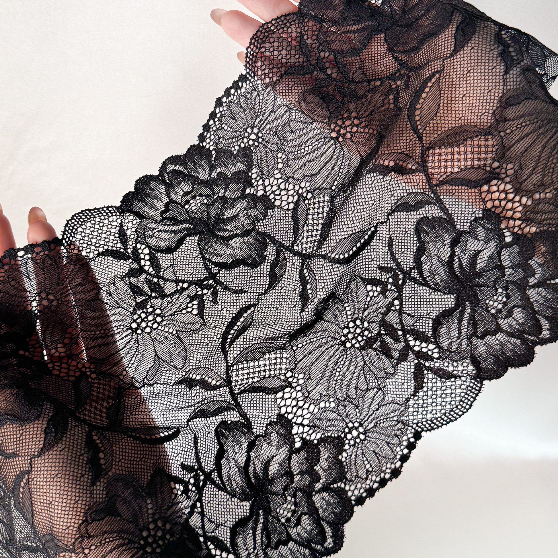 Hands showcasing black elastic lace fabric, 21 cm wide, highlighting its flexibility and elegant floral details, ideal for lingerie projects.