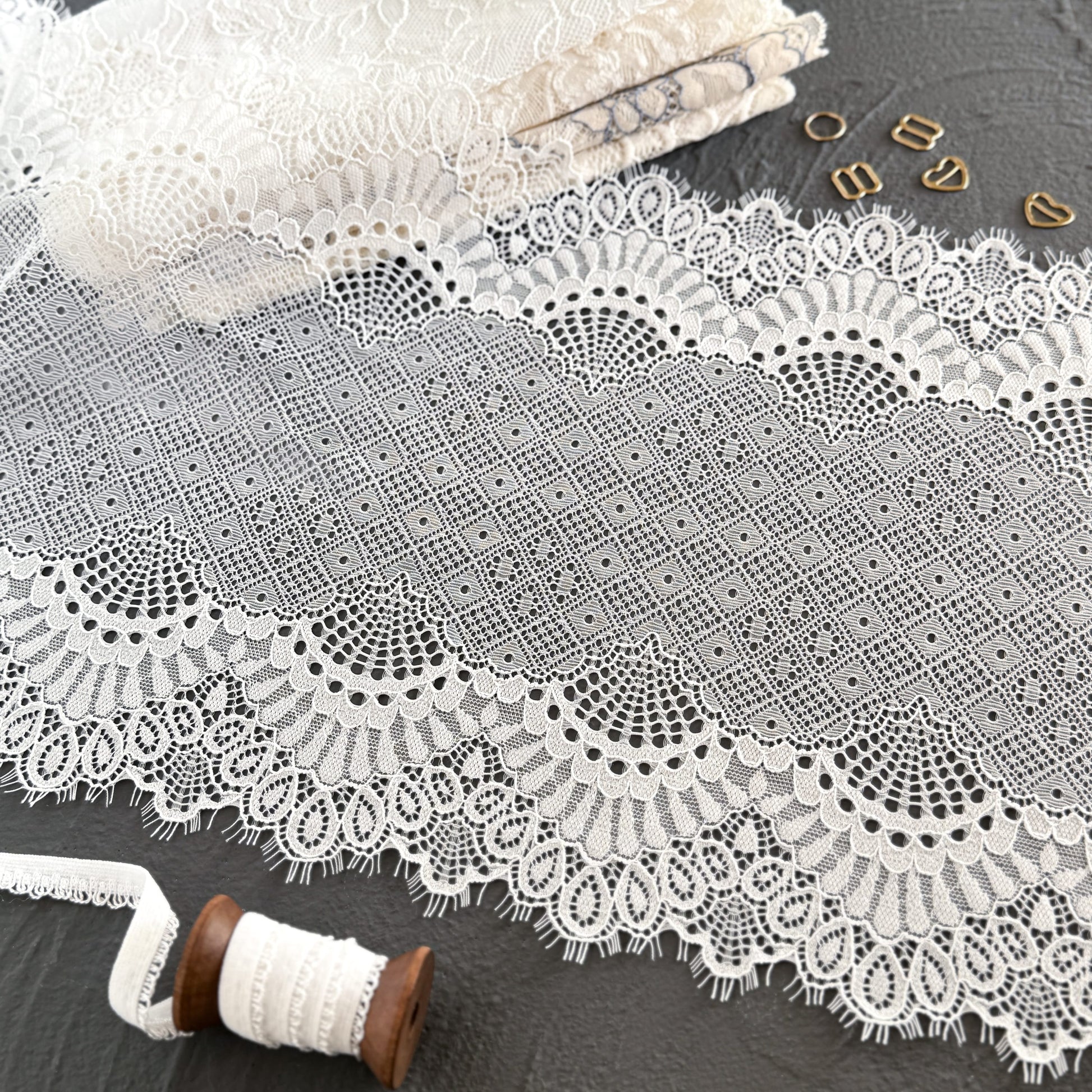 Wide off-white elastic lace trim with delicate eyelashes, 23 cm wide, featuring intricate geometric and scalloped patterns, styled with sewing accessories on a dark gray surface.