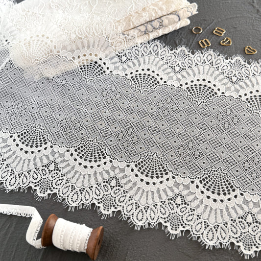 Wide off-white elastic lace trim with delicate eyelashes, 23 cm wide, featuring intricate geometric and scalloped patterns, styled with sewing accessories on a dark gray surface.