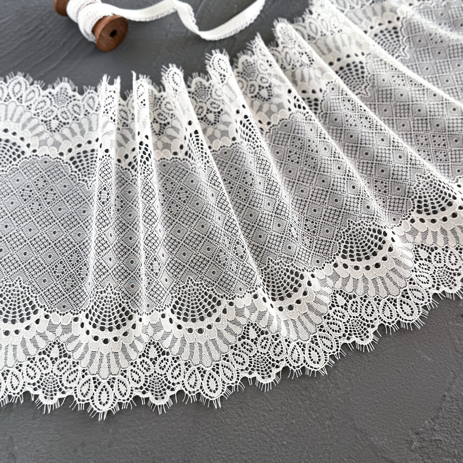 Close-up of off-white elastic lace with eyelash edges, 23 cm wide, showcasing its detailed geometric and scalloped designs, ideal for lingerie or garment accents.