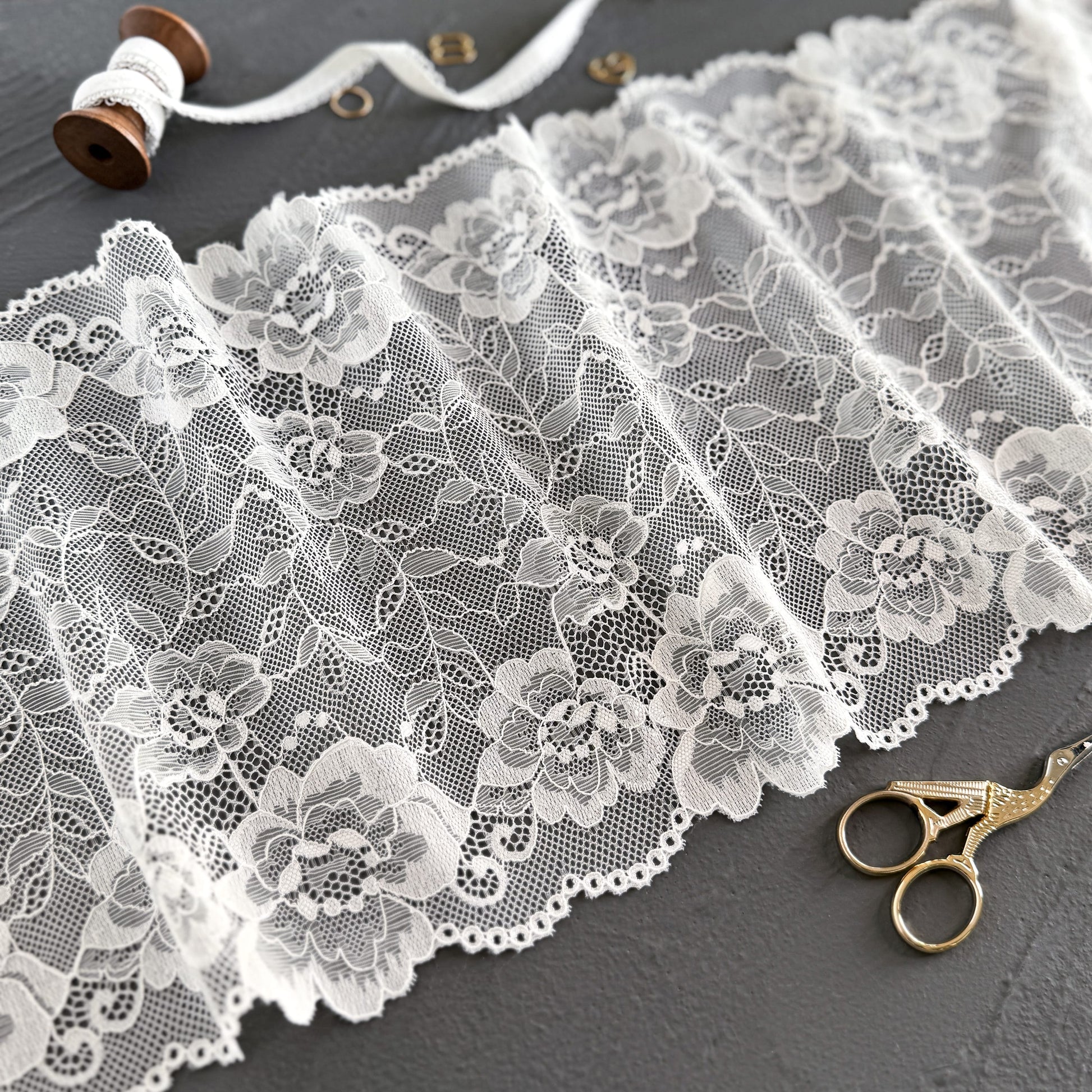 Off-white elastic lace fabric, 23 cm wide, featuring intricate floral patterns, styled with sewing accessories on a dark gray background for lingerie-making inspiration.