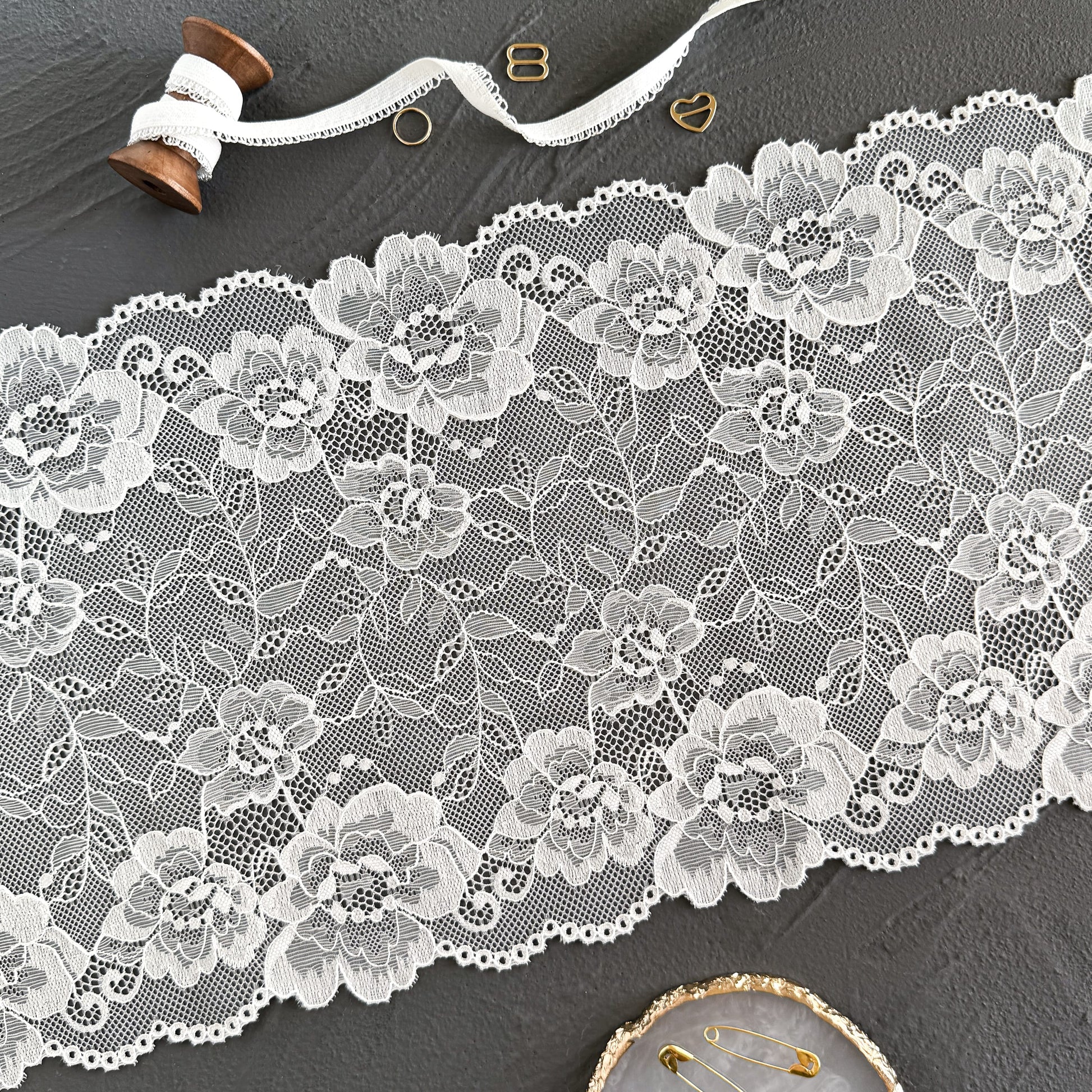 Off-white elastic lace fabric, 23 cm wide, laid flat to highlight its floral patterns and delicate craftsmanship, ideal for intimate apparel designs.