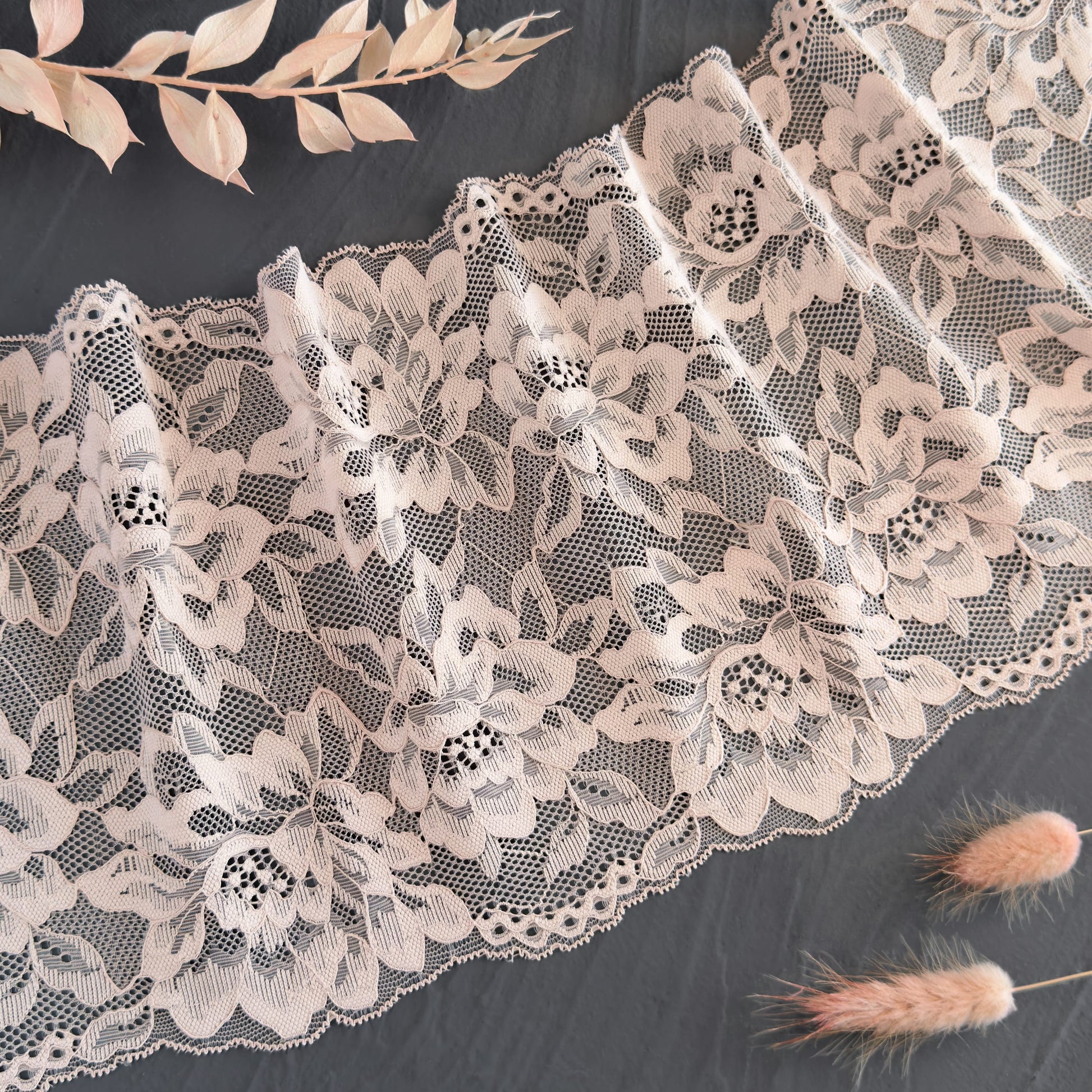 Beige elastic lace fabric, 21 cm wide, featuring delicate floral patterns, styled with decorative dried foliage, perfect for lingerie making.